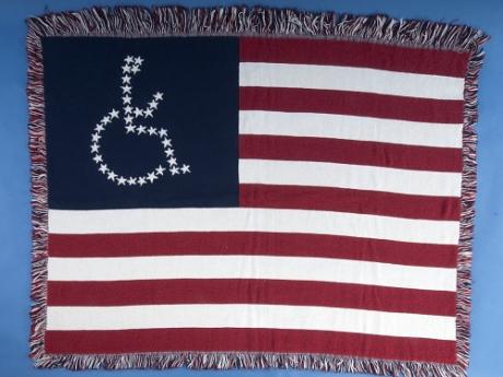 An American flag woven lap blanket. Instead of the 50 white stars making up the upper left corner of the flag, the stars are arranged in the shape of the international symbol for accessibility- a figure in a wheelchair. 
